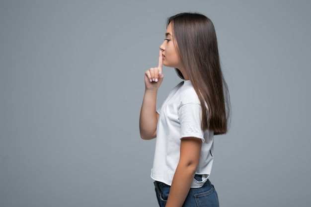 Ritratto laterale di bella donna asiatica che sorride e che mostra le mani del segno per zitto su fondo grigio