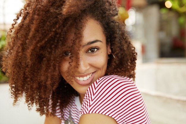 Ritratto laterale di donna sorridente felice con l'acconciatura riccia folta vestita casualmente, pensa a qualcosa di piacevole, si gode il riposo estivo nel paese del resort. Felicità ed emozioni positive.