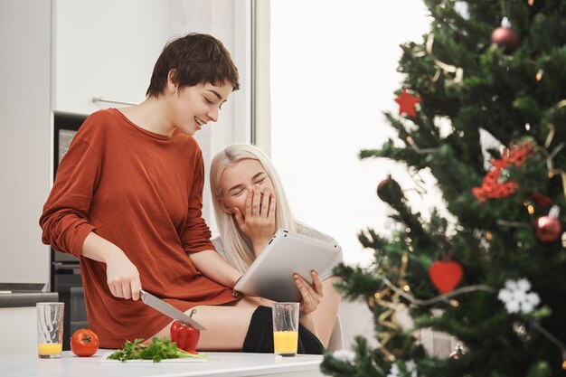 Ritratto laterale di un'attraente ragazza magra con i capelli a camicia che taglia le verdure e mostra qualcosa in tavoletta per far arrabbiare un amico che ride ad alta voce e si diverte a passare del tempo con lei