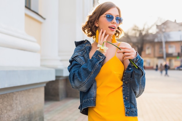 Ritratto luminoso di bella donna che tiene fiore, vestito giallo, giacca di jeans, stile hipster, tendenza moda estiva, sorriso, occhiali da sole alla moda