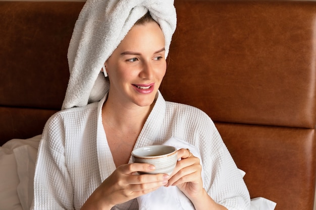 Ritratto mattutino di una bella donna che si rilassa a letto dopo la doccia indossando accappatoio e asciugamano in testa, bevendo un gustoso tè