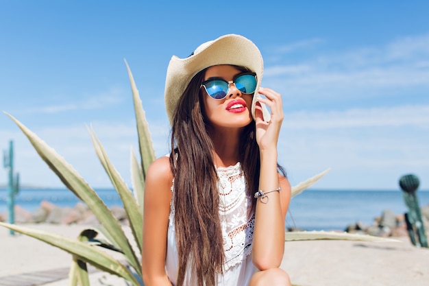 Ritratto orizzontale del primo piano della ragazza attraente del brunette con capelli lunghi che si siede sulla spiaggia vicino al cactus sui precedenti. Sta sorridendo lontano.