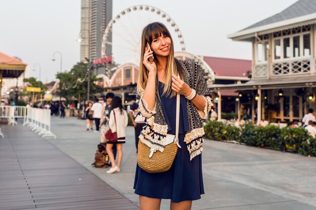 Ritratto positivo di estate della donna allegra in vestito alla moda che parla dal telefono cellulare e che sorride sul lungofiume a Bangkok