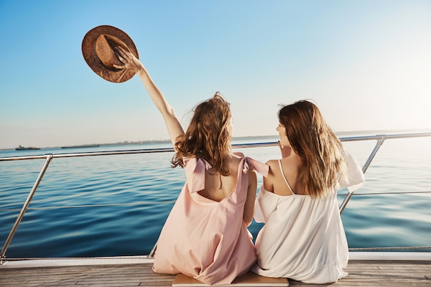 Ritratto posteriore di due amiche seduta sulla barca, agitando con cappello mentre parla e godendo guardando in riva al mare. Alla fine le sorelle si sono messe in vacanza per visitare la loro mamma che vive in Italia