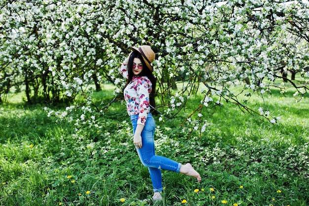 Ritratto primaverile di una ragazza bruna con occhiali rosa e cappello al giardino di fiori verdi