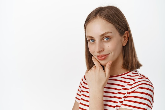 Ritratto ravvicinato di bella giovane donna con capelli biondi, occhi azzurri e pelle luminosa naturale senza trucco, viso pulito, toccare il mento e sguardo premuroso, sorriso sicuro di sé, sfondo bianco