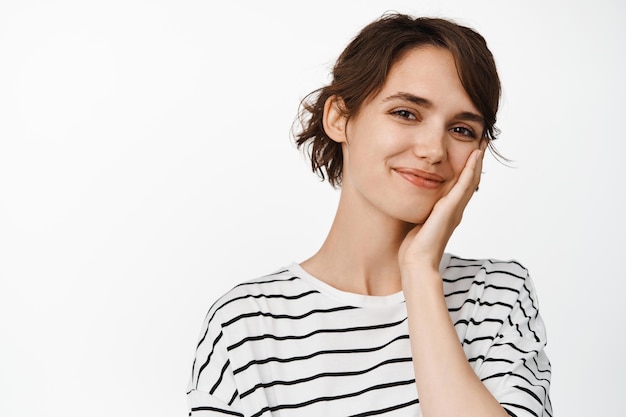 Ritratto ravvicinato di bella giovane donna sorridente, toccando una pelle sana e pulita, pubblicità di cosmetici per la cura della pelle, in piedi su sfondo bianco