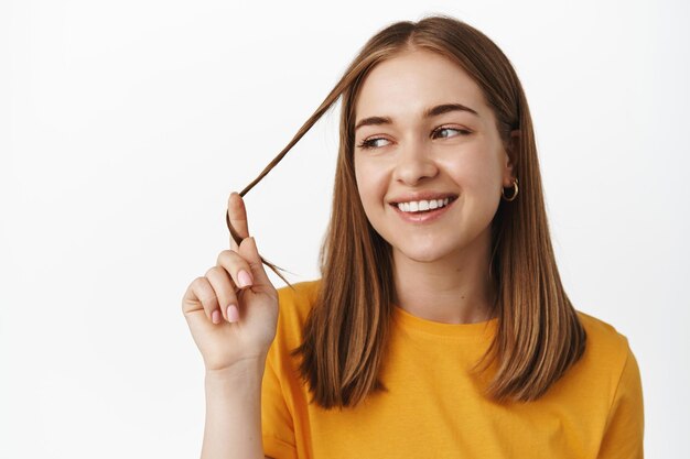 Ritratto ravvicinato di bella ragazza che gioca con una ciocca di capelli, sorridente e che guarda da parte rilassata, posa in maglietta gialla, espressione spensierata, sfondo bianco