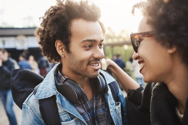 Ritratto ravvicinato di coppia afro-americana in abiti alla moda e tagli di capelli afro, abbracciati sorridendo e guardandosi l'un l'altro, in piedi nel parco. Coppia innamorata che trascorre i fine settimana all'aperto.