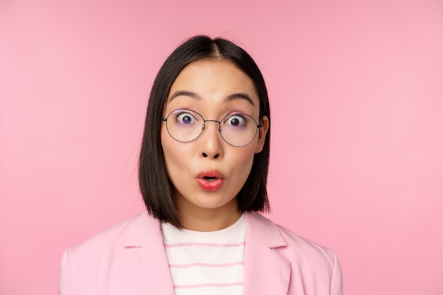 Ritratto ravvicinato di donna d'affari asiatica con gli occhiali che guarda sorpreso dalla reazione stupita della fotocamera in piedi in tuta su sfondo rosa