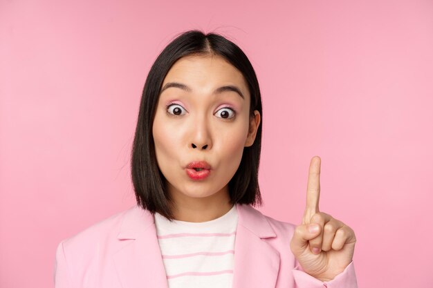Ritratto ravvicinato di donna d'affari che alza il dito suggerendo di avere un'idea o una soluzione in piedi su sfondo rosa
