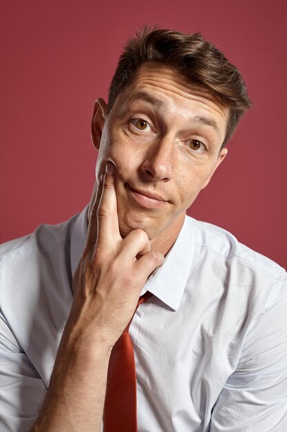 Ritratto ravvicinato di un interessante maschio brunet con occhi marroni, che indossa una camicia bianca e una cravatta di terracotta. Sta guardando la telecamera mentre posa in uno studio su uno sfondo rosso. Concetto o