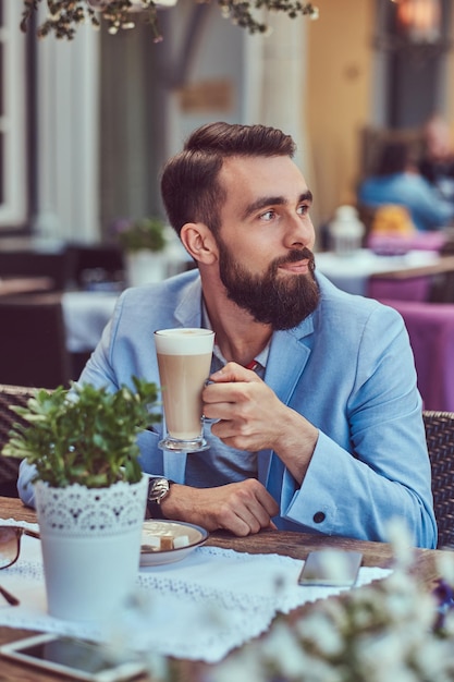 Ritratto ravvicinato di un maschio barbuto alla moda con un taglio di capelli elegante, beve un bicchiere di cappuccino, seduto in un caffè all'aperto.