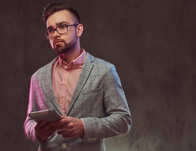 Ritratto ravvicinato di un uomo barbuto sicuro ed elegante con acconciatura e occhiali da sole in un abito grigio e camicia rosa, tiene un tablet in uno studio. Isolato su sfondo grigio.