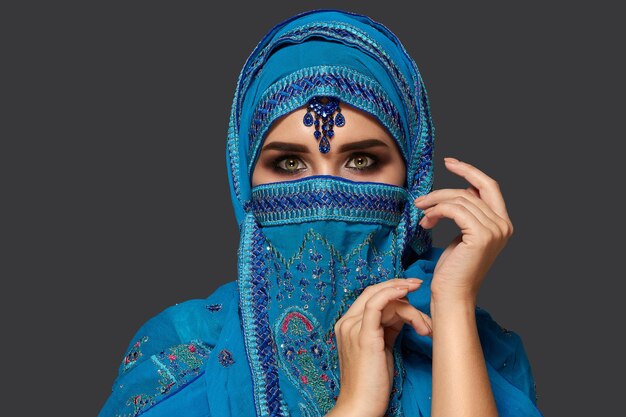Ritratto ravvicinato di una bella donna con bellissimi occhi fumosi che indossa un hijab blu decorato con paillettes e gioielli. Sta gesticolando e guardando la telecamera su uno sfondo scuro. Emozione umana