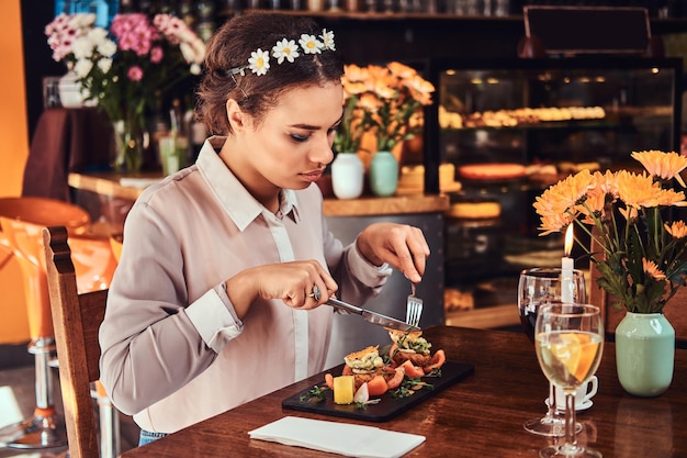 Ritratto ravvicinato di una bella donna dalla pelle nera che indossa una camicetta e una fascia di fiori, godendosi la cena mentre si mangia in un ristorante.