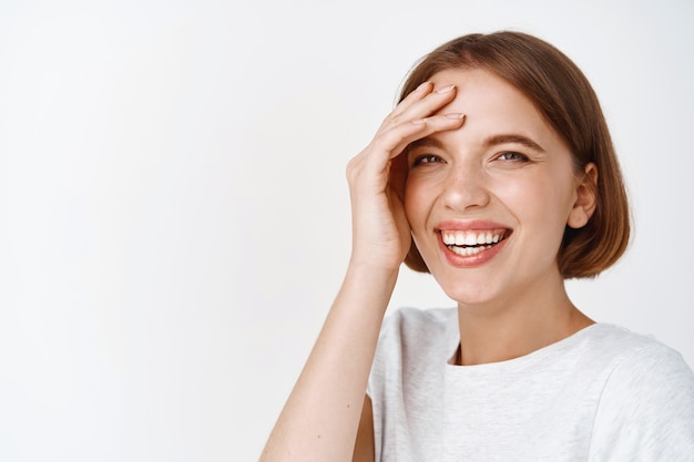 Ritratto ravvicinato di una bellissima modella femminile con i capelli corti, sorridente e ridente spensierata, toccando il viso eccitato, in piedi contro il muro bianco