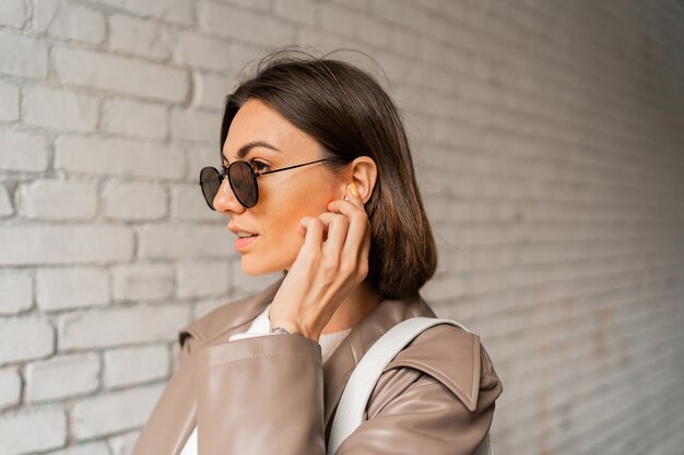 Ritratto ravvicinato di una donna elegante dai capelli corti con auricolari in cappotto di pelle casual e occhiali da sole in posa su un muro di mattoni urbano