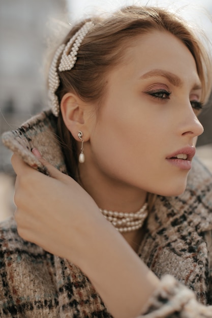 Ritratto ravvicinato di una giovane donna bionda graziosa in gioielli di perle e cappotto di tweed a scacchi in posa all'aperto