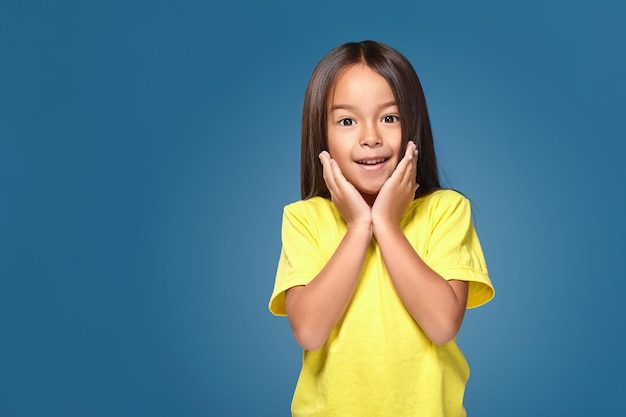 Ritratto ravvicinato di una graziosa bambina gioiosa carina con una pelle eccellente e un sorriso raggiante, sta ammirando la sua bellezza in uno specchio, su sfondo blu