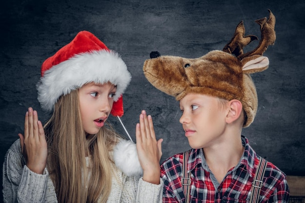 Ritratto ravvicinato di una ragazza bionda con il cappello di Babbo Natale e un ragazzo carino con un cappello di cervo divertente su sfondo grigio.