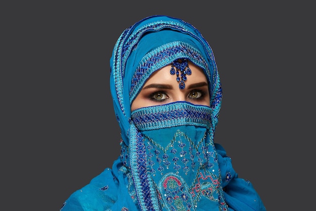 Ritratto ravvicinato di una splendida giovane donna con bellissimi occhi fumosi che indossa un elegante hijab blu decorato con paillettes e gioielli. Sta posando di lato e guardando la telecamera su uno sfondo scuro