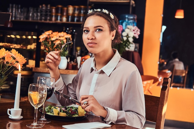 Ritratto ravvicinato felice di una bella donna dalla pelle nera che indossa una camicetta e una fascia di fiori, godendosi la cena mentre si mangia in un ristorante.