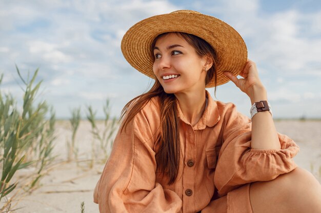 Ritratto romantico della donna sorridente in cappello di paglia e vestito di lino alla moda. Ragazza vaga agghiacciante vicino all'oceano. Tendenza moda estiva.