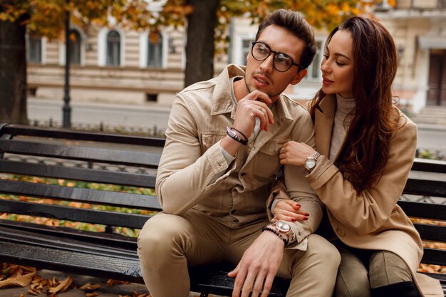 Ritratto romantico di giovani belle coppie nell'amore che abbraccia e che bacia sul banco nel parco di autunno. Indossa un elegante cappotto beige.
