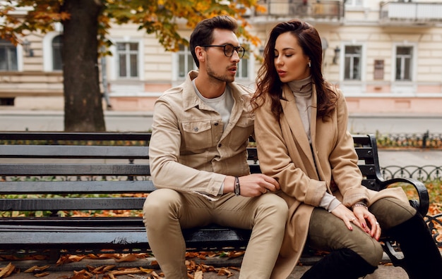 Ritratto romantico di giovani belle coppie nell'amore che abbraccia e che bacia sul banco nel parco di autunno. Indossa un elegante cappotto beige.