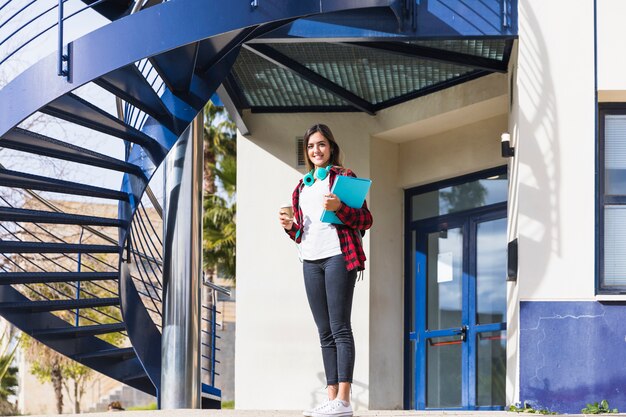 Ritratto sorridente dei libri della tenuta di una giovane donna e della tazza di caffè asportabile che stanno davanti all&#39;edificio universitario