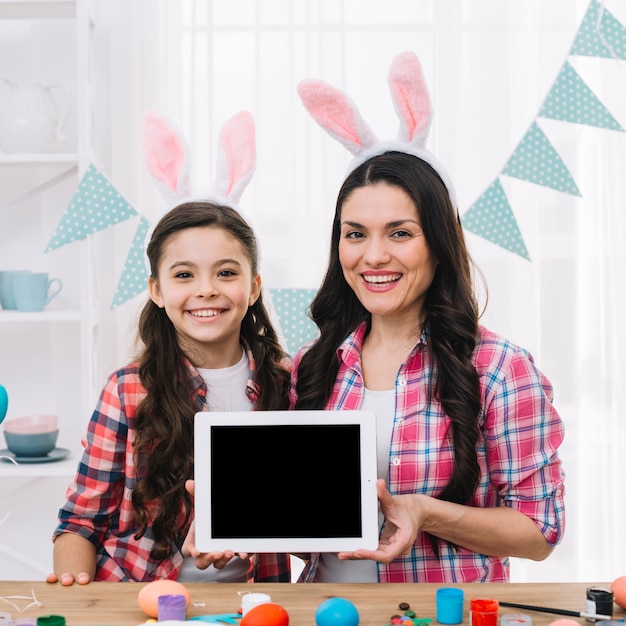 Ritratto sorridente della madre e della figlia che mostrano compressa digitale dietro la tavola di legno con le uova di Pasqua