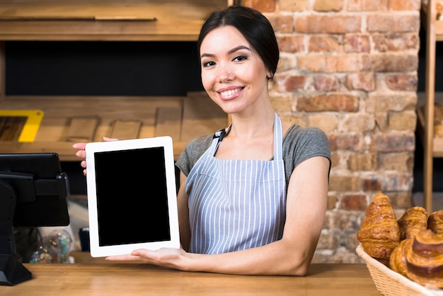 Ritratto sorridente di giovane panettiere femminile che tiene compressa digitale che sta al contatore del forno