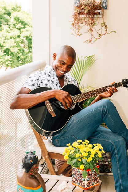 Ritratto sorridente di un giovane africano che si siede sulla sedia che gioca la chitarra nel balcone