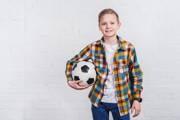 Ritratto sorridente di un pallone da calcio della tenuta del ragazzo che sta in piedi contro il muro di mattoni bianco