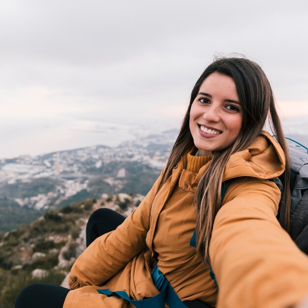 Ritratto sorridente di una giovane viandante femminile che prende selfie che si siede sulla cima della montagna