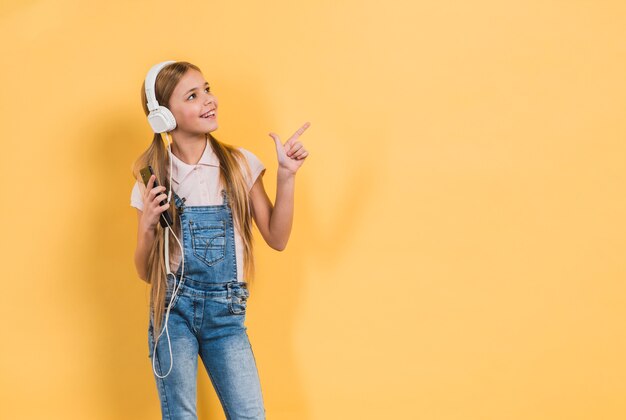 Ritratto sorridente di una musica d&#39;ascolto della ragazza sulla cuffia che indica a qualcosa contro fondo giallo