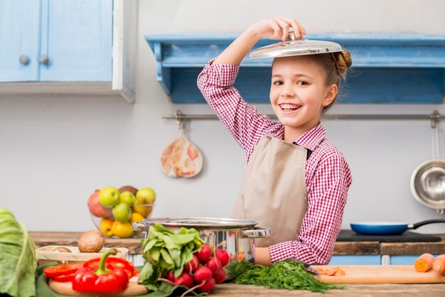 Ritratto sorridente di una ragazza con il coperchio sopra la sua testa in piedi in cucina