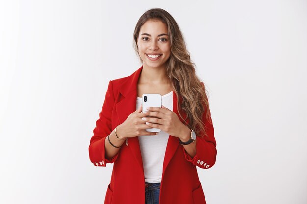 Ritratto splendida donna alla moda sicura di sé che indossa una giacca rossa chiedendo di scattare una foto tenendo lo smartphone sorridente ampiamente come prendere selfie specchio guardando bene vestito, postare immagini online