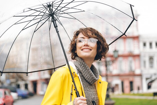 Ritratto stupefacente della donna felice in impermeabile giallo che cammina nella città sotto l'ombrello trasparente durante il giorno piovoso freddo