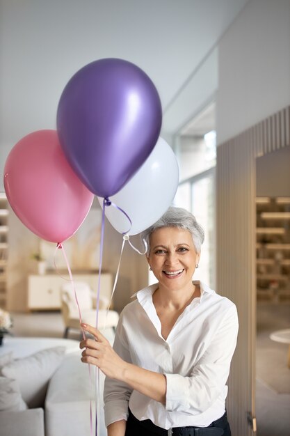 Ritratto verticale della donna matura dai capelli grigi alla moda in camicia bianca che celebra il successo, la vittoria o l'anniversario