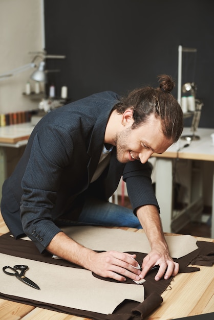 Ritratto verticale di allegro attraente caucasico maturo stilista maschio con acconciatura alla moda in abito nero, lavorando su una nuova collezione di abiti per sfilata di moda, tagliando le parti del vestito