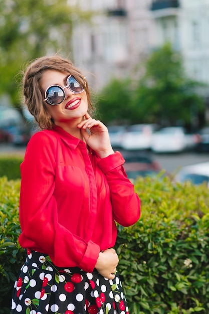 Ritratto verticale di bella ragazza in occhiali da sole in posa per la telecamera nel parco. Indossa una camicetta rossa, una gonna nera e una bella acconciatura. Sta sorridendo alla telecamera.