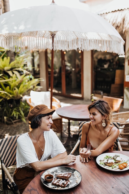 Ritratto verticale di donne abbronzate in abiti alla moda che sorridono e parlano nell'accogliente street cafe