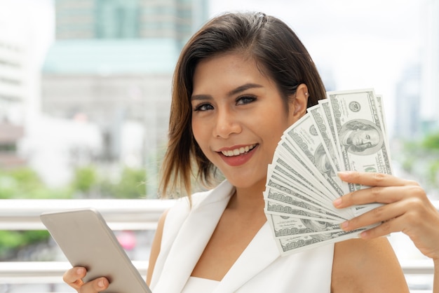 Riuscita bella giovane donna asiatica di affari che usando le fatture del dollaro americano dei soldi e dello Smart Phone a disposizione