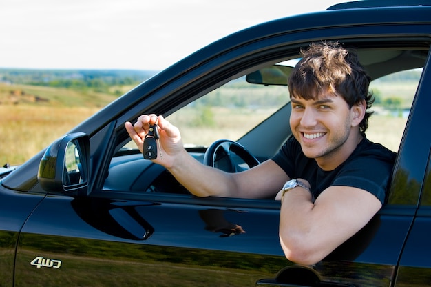 Riuscito giovane uomo felice che mostra le chiavi che si siedono nella nuova automobile