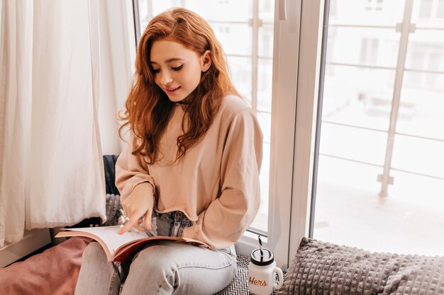 Rivista attraente della lettura della ragazza dai capelli lunghi. Ritratto interno di accattivante signora allo zenzero con libro.