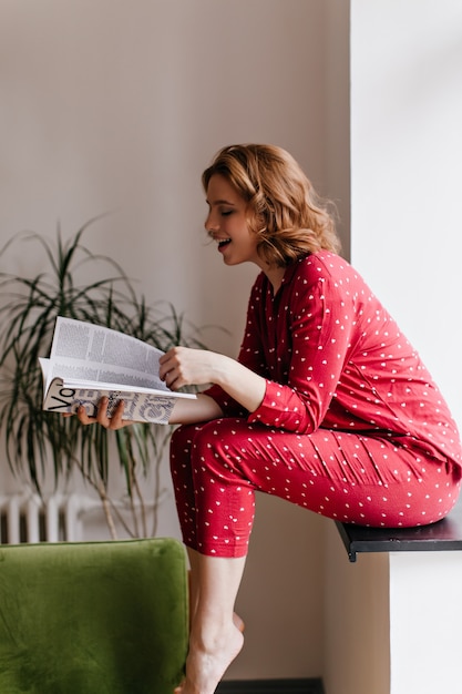 Rivista di lettura della donna a piedi nudi di risata mattina. Tiro al coperto di bella donna in pigiama seduto sul davanzale della finestra.