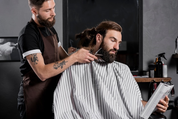 Rivista maschio della lettura del cliente mentre tagliando la sua barba