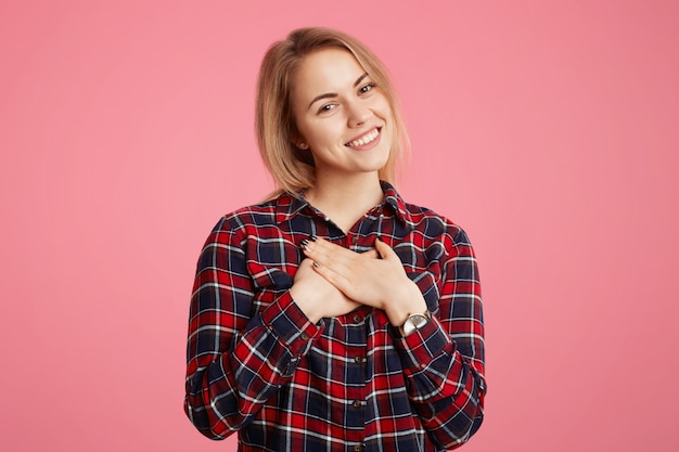 Romantica bella giovane femmina con piacevole sorriso splendente, tiene le mani sul petto